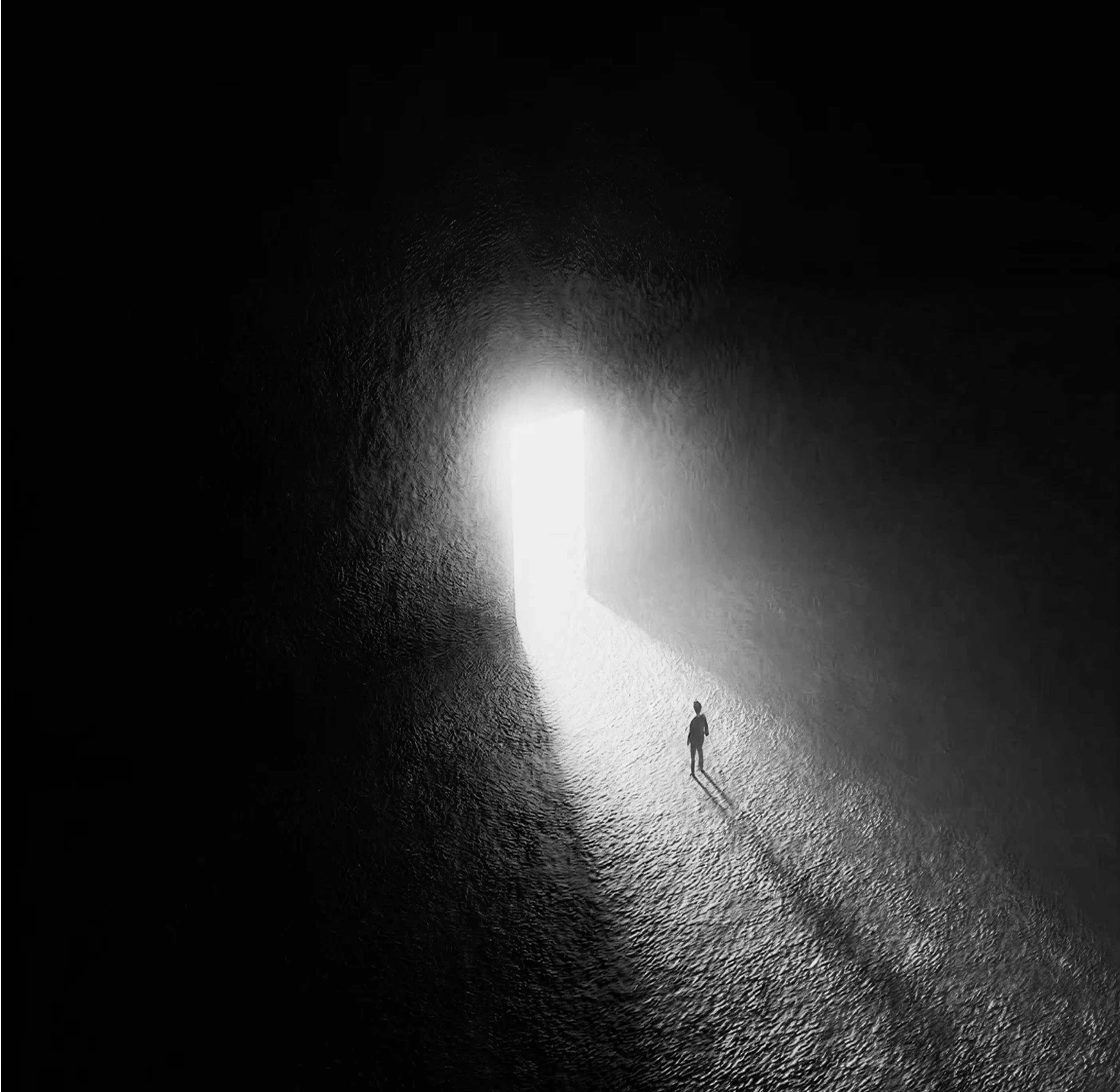 Black-and-white overhead view of a lone figure walking out of a lit cave mouth onto a bright path.