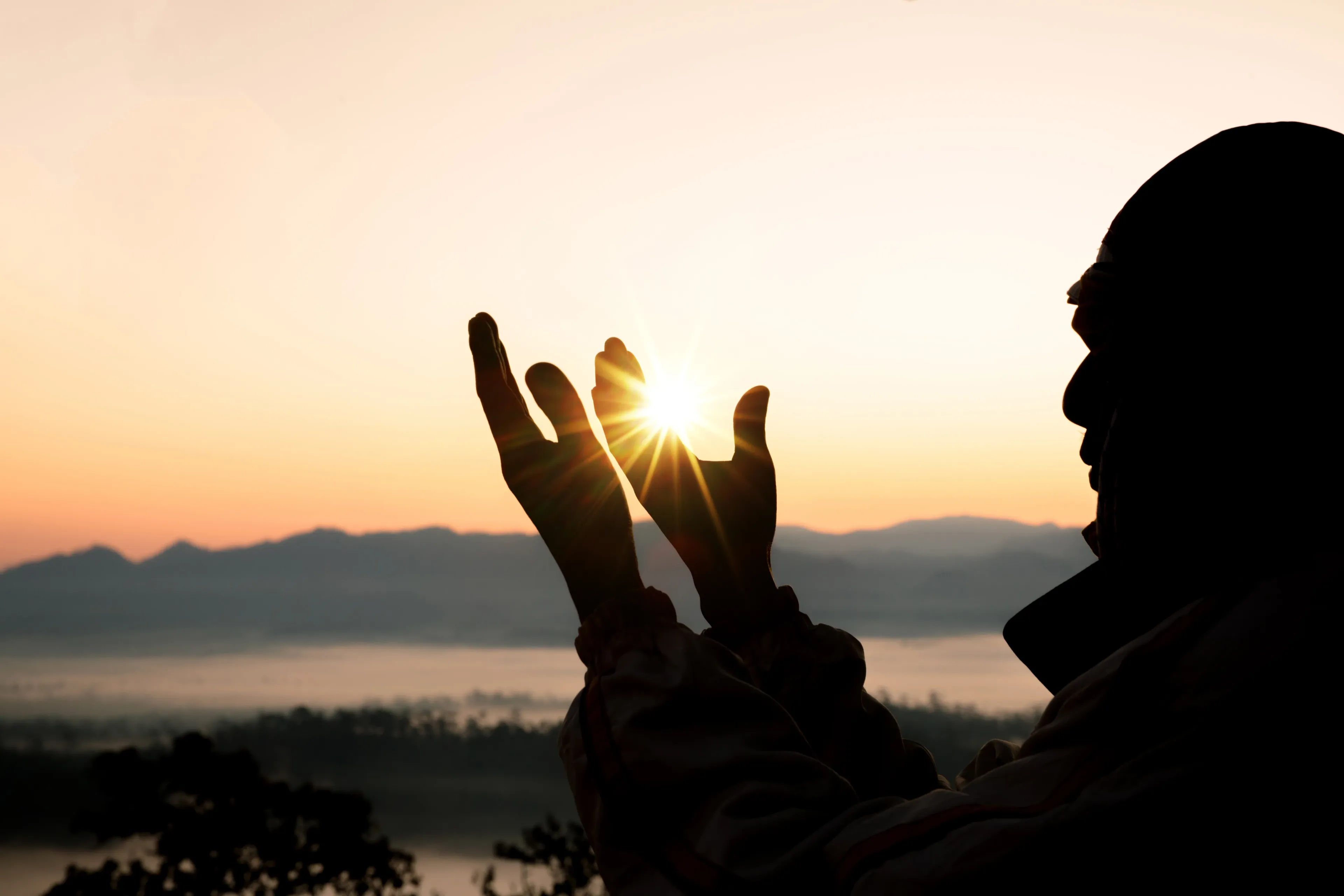 Cupped hands are raised in du'a, silhouetted against a sunrise sky.