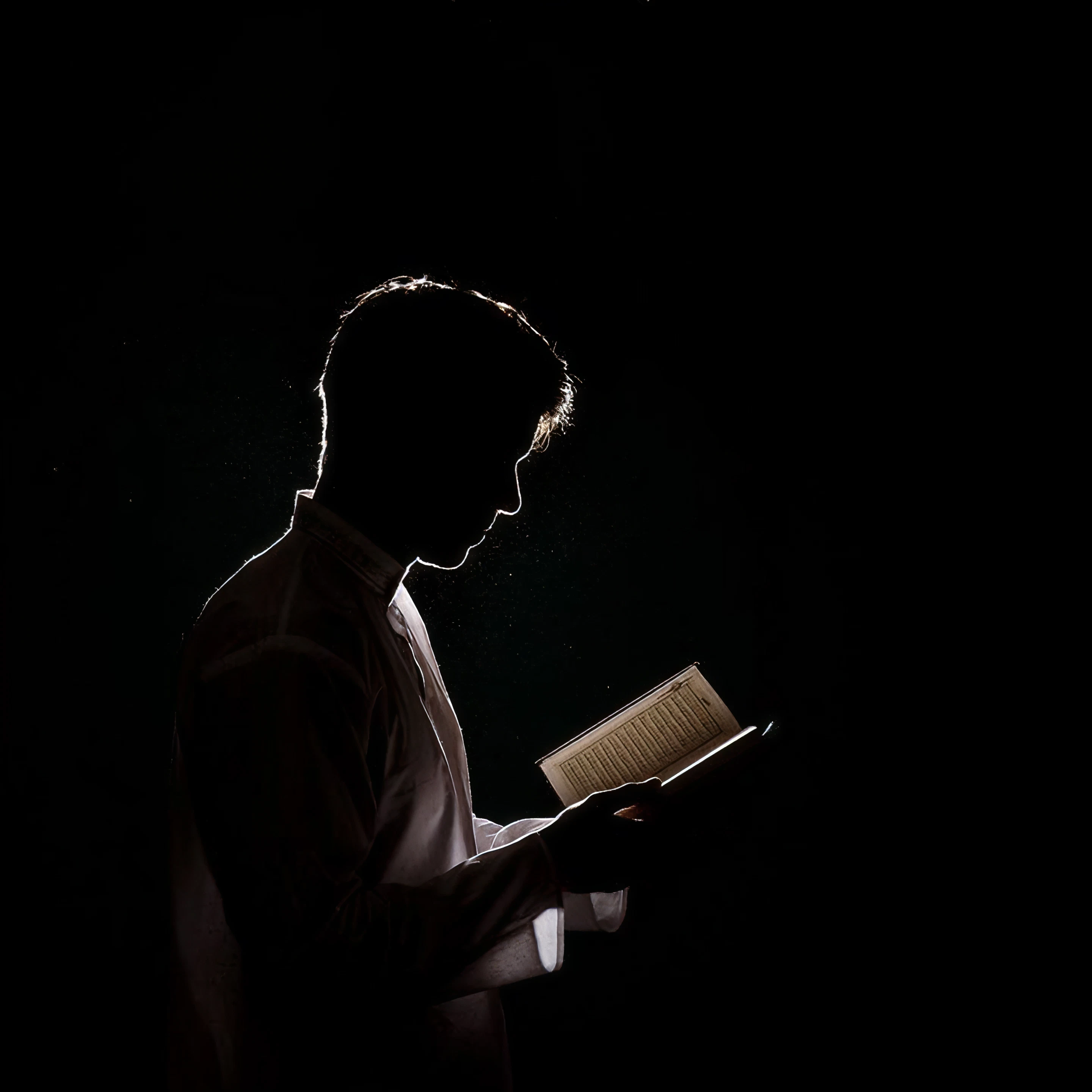 A man in white robes reads from an open Qur'an, rim-lit against a black background.