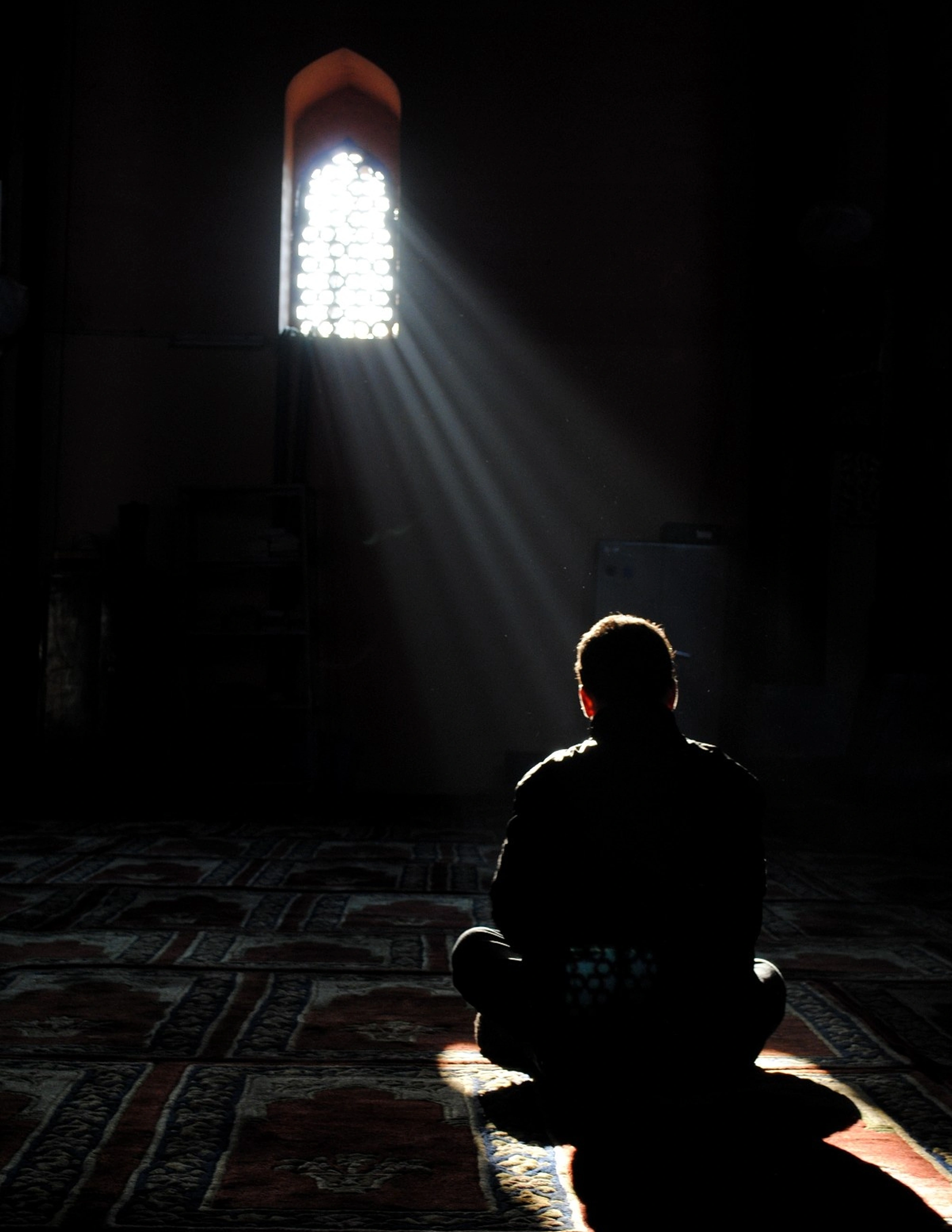 A man sits with his back to camera on a red patterned prayer carpet in a near-black mosque interior, in silhouette under hard vertical rays from one small pierced-screen window.