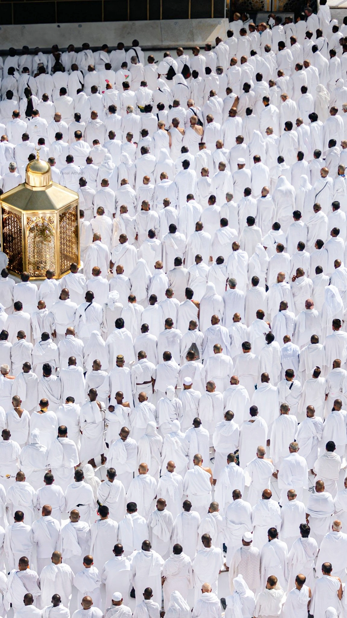 Overhead view of a dense crowd of worshippers in white ihram garments standing in rows, with a gold lantern at the edge of the courtyard.
