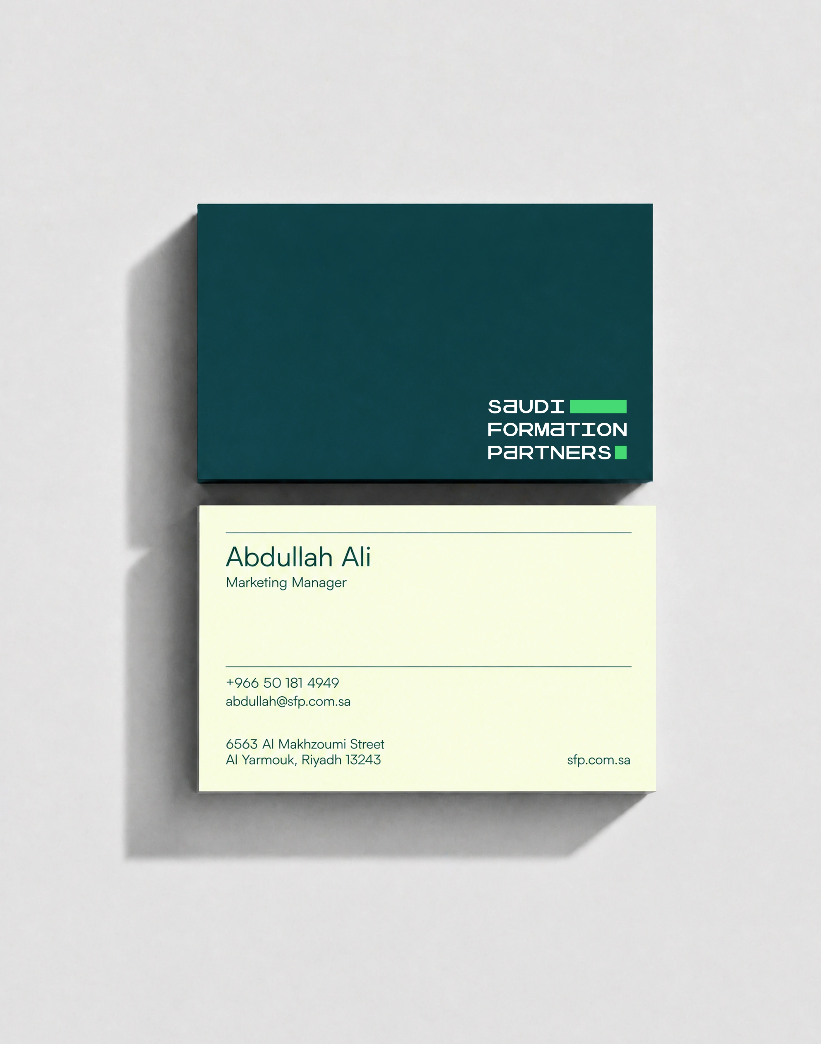 A pair of business cards photographed flat from above on a light grey surface, the dark teal back carrying the wordmark resting on the cream front, which shows a name, job title, phone number, email address and a Riyadh street address.