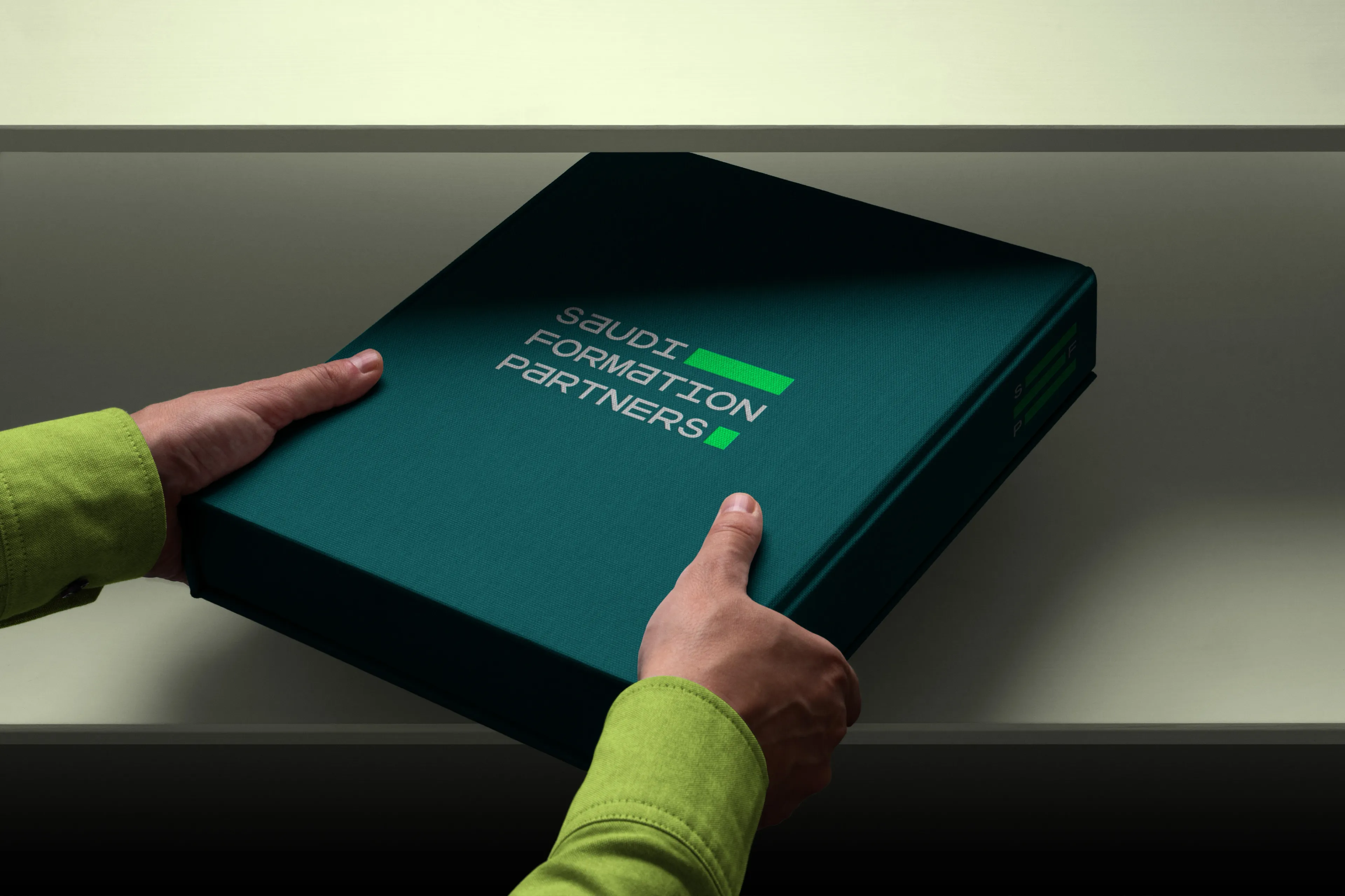 A hand in a lime-green sleeve lifts the lid from a dark teal cloth presentation box printed with the Saudi Formation Partners wordmark.