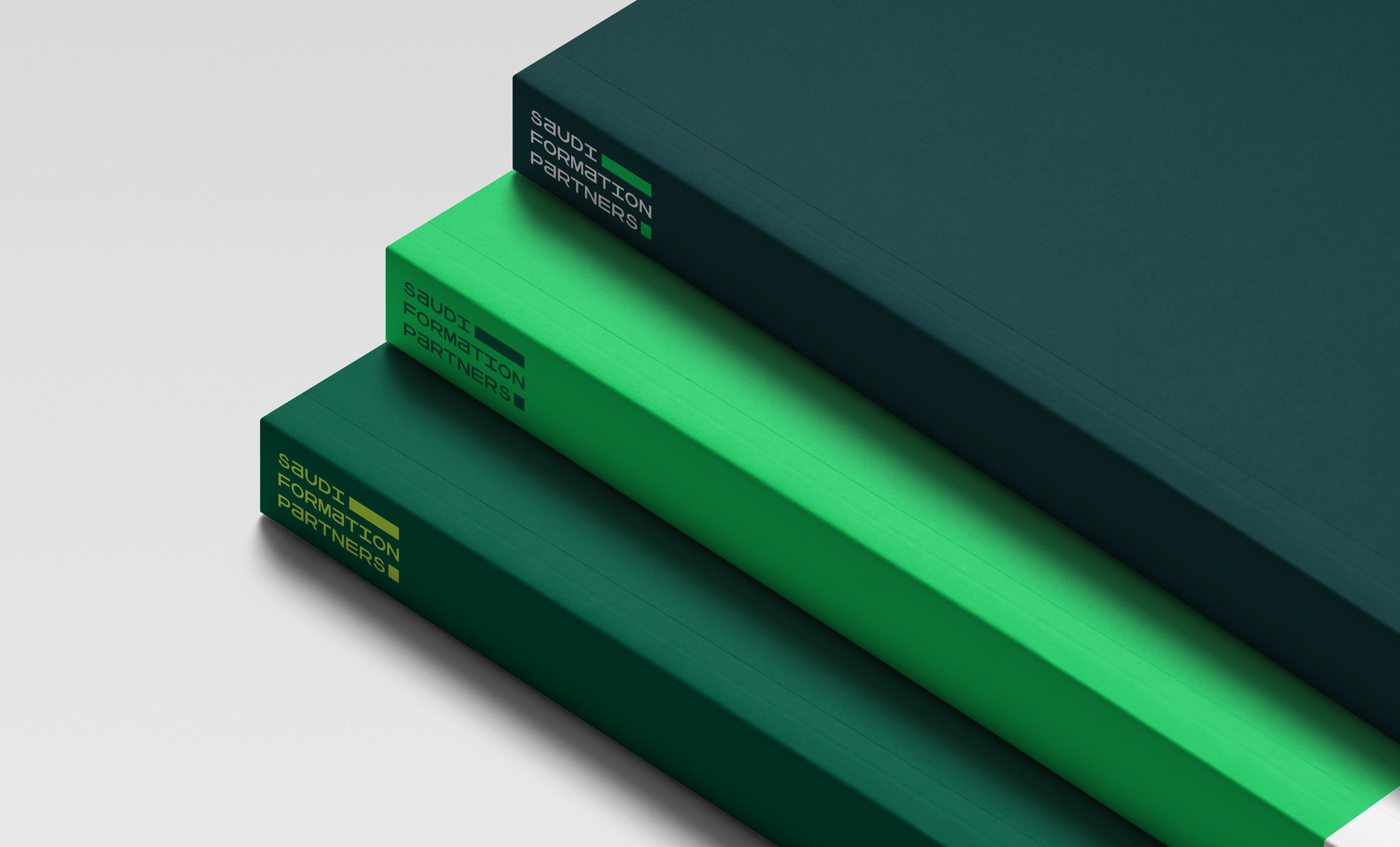 Three branded slipcase boxes in dark green, malachite green and Tiber green stacked so each steps back from the one below, on a pale grey ground, every spine carrying the rotated Saudi Formation Partners wordmark.