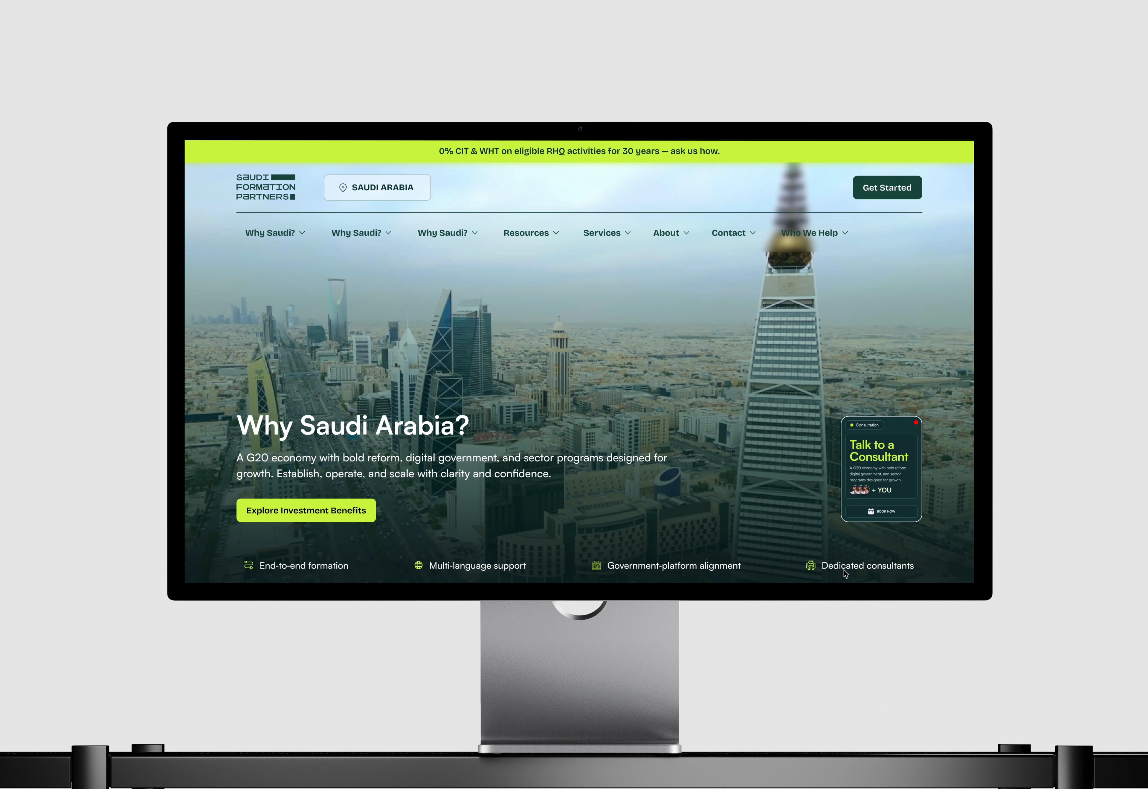 The Saudi Formation Partners website displayed on a studio desktop monitor, a lime announcement bar above the header and an aerial Riyadh skyline hero headed Why Saudi Arabia, with a lime Explore Investment Benefits button and a floating Talk to a Consultant card at the right.