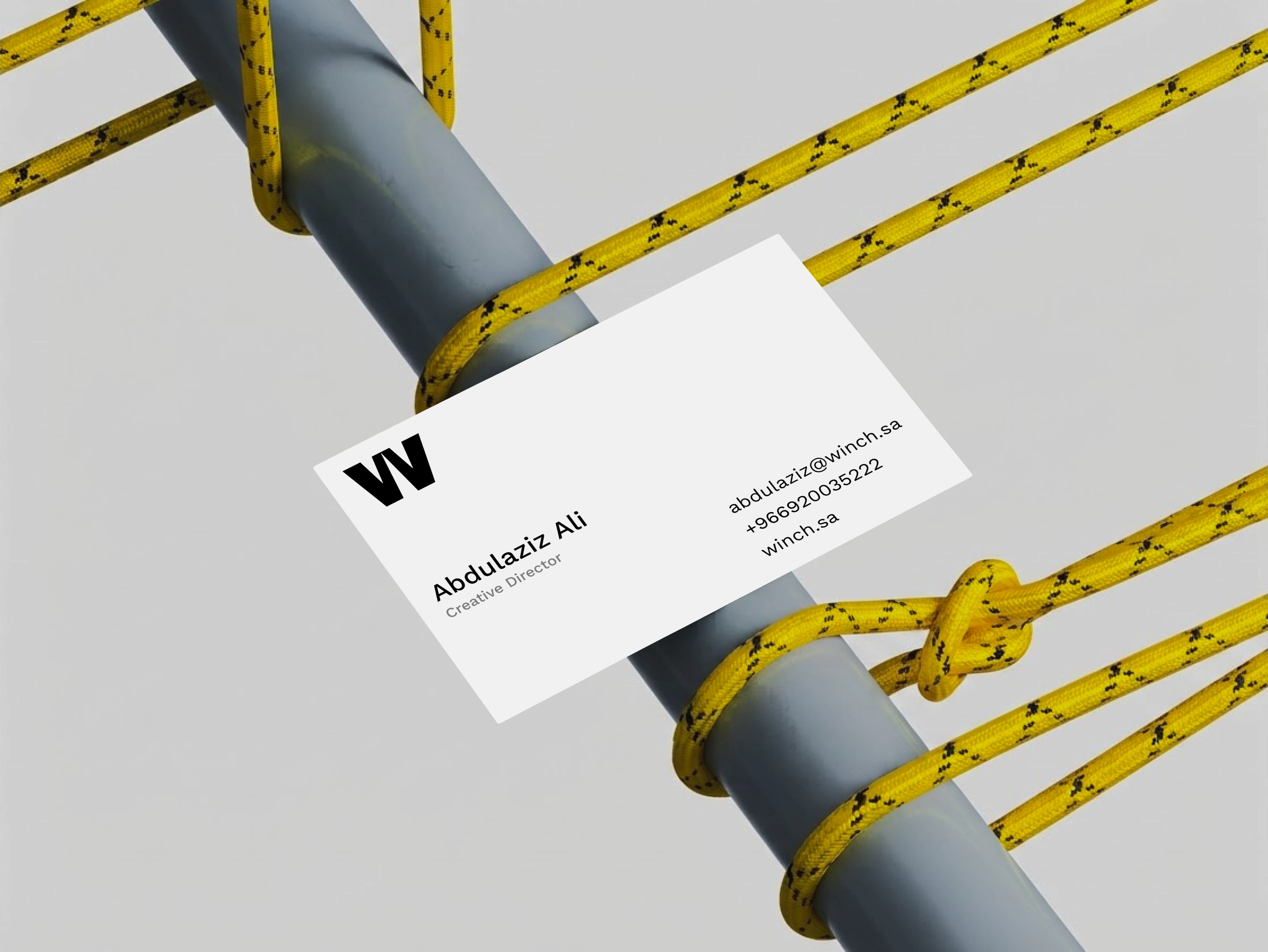 A single white WINCH business card wedged against a grey pipe criss-crossed by knotted yellow rope, printed with the name Abdulaziz Ali, the title Creative Director, an email address, a phone number and winch.sa.