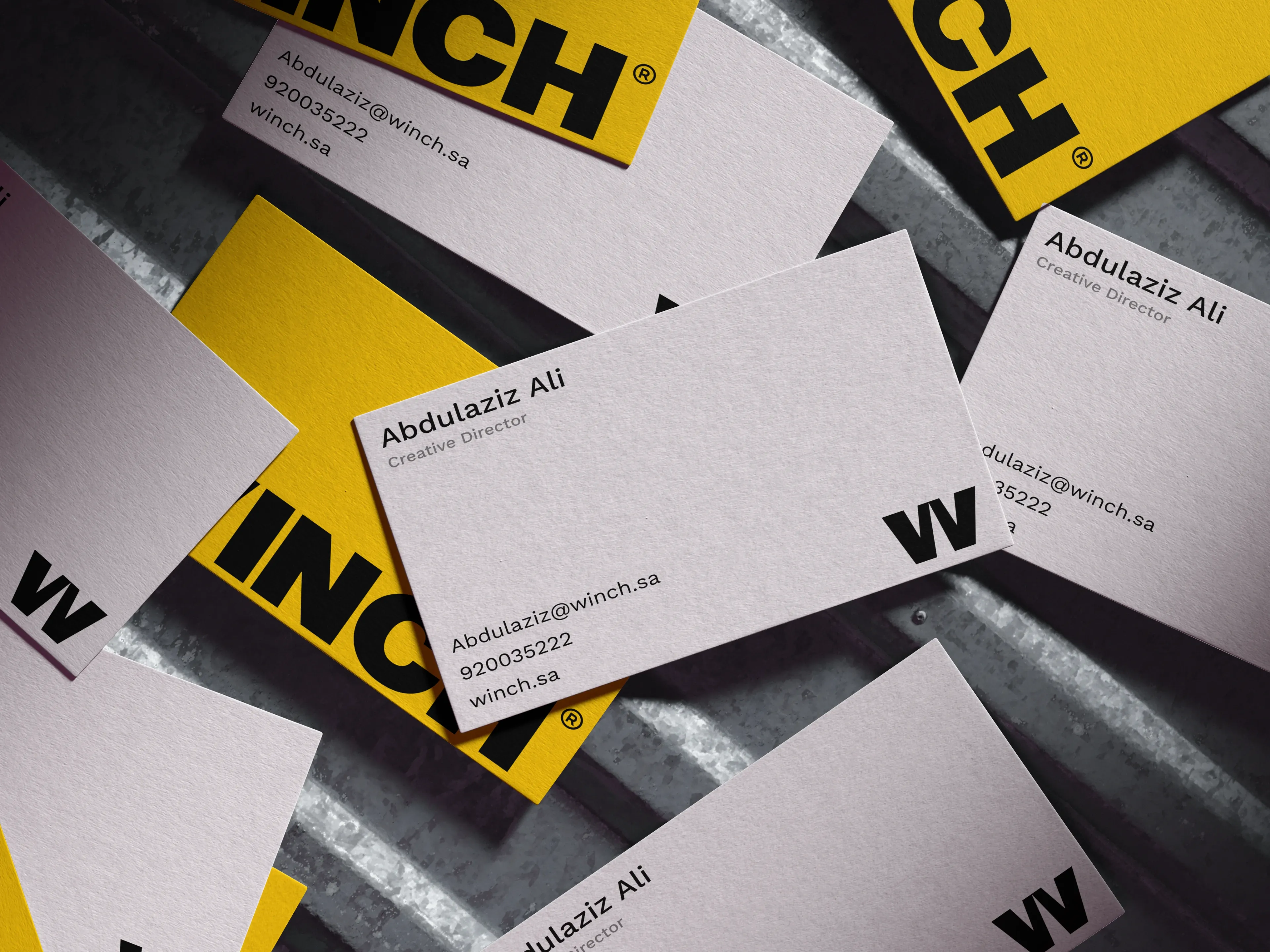 White and yellow WINCH business cards scattered across a brushed metal surface, the card in focus showing the W monogram above a name, job title, email address and phone number.