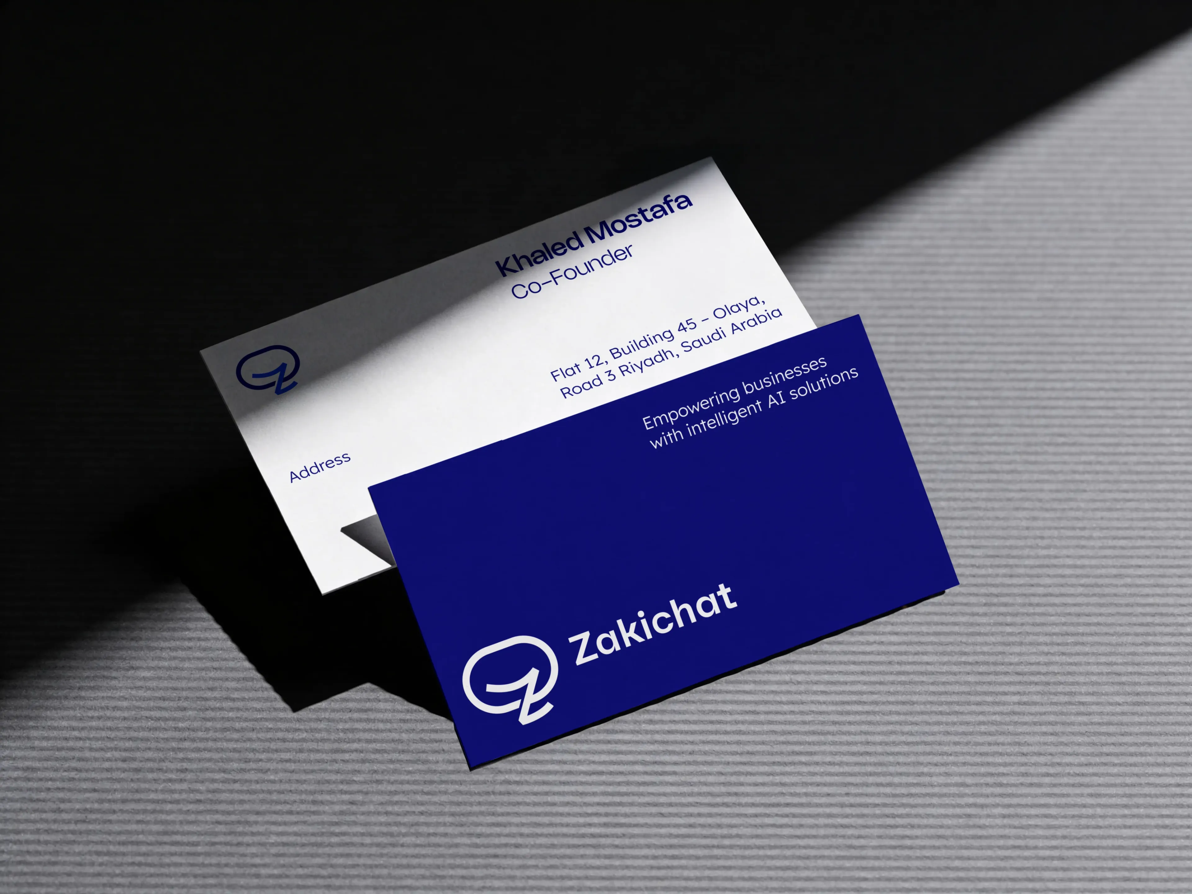 A white and a navy Zakichat business card lying apart at different angles and partly overlapping, the white card carrying the name and a Riyadh address and the navy card the lockup with the line Empowering businesses with intelligent AI solutions.
