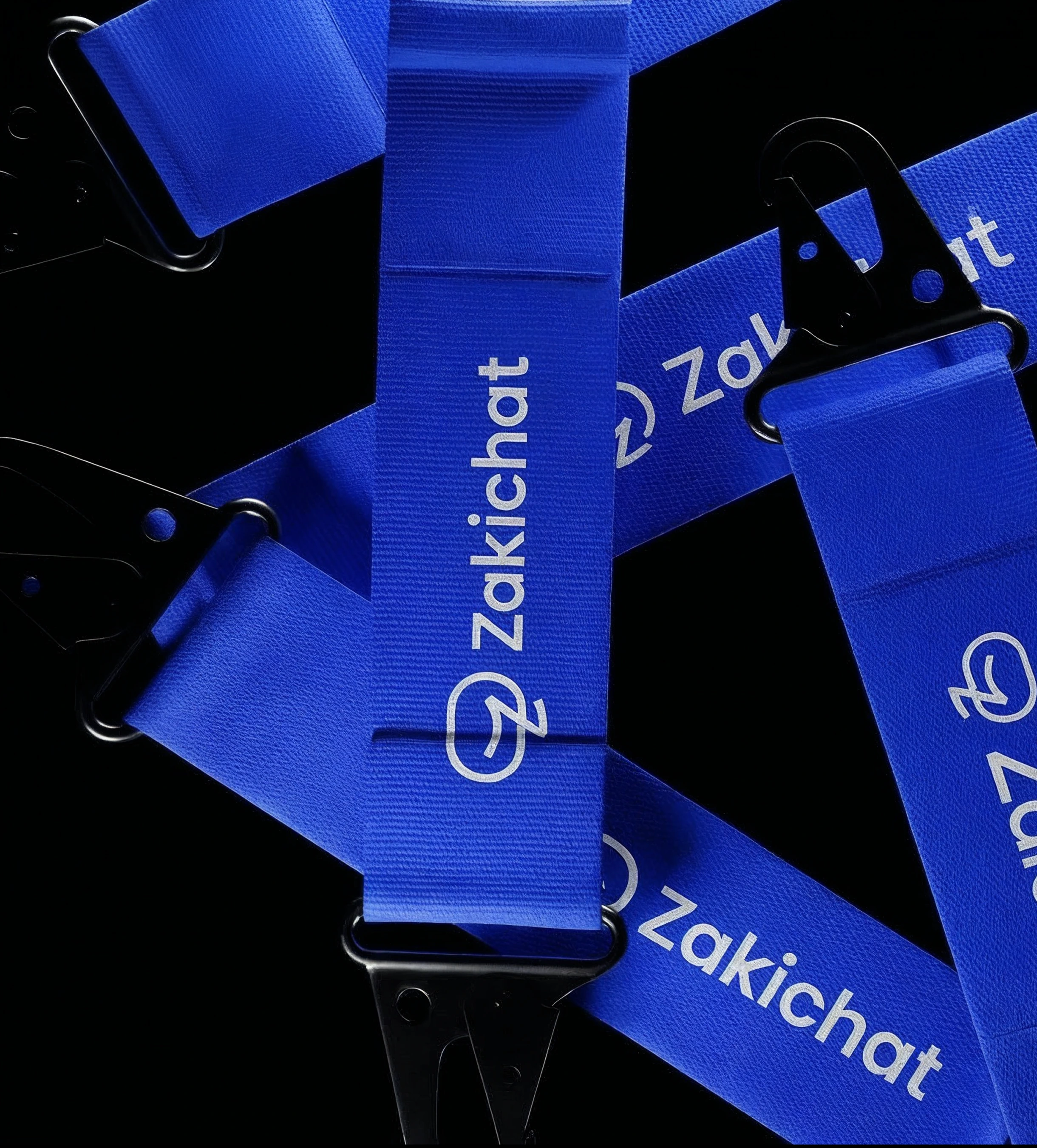 A pile of blue woven Zakichat lanyard straps crossing a near-black ground at angles, printed with the white mark and wordmark and fitted with black D-rings and swivel clips.