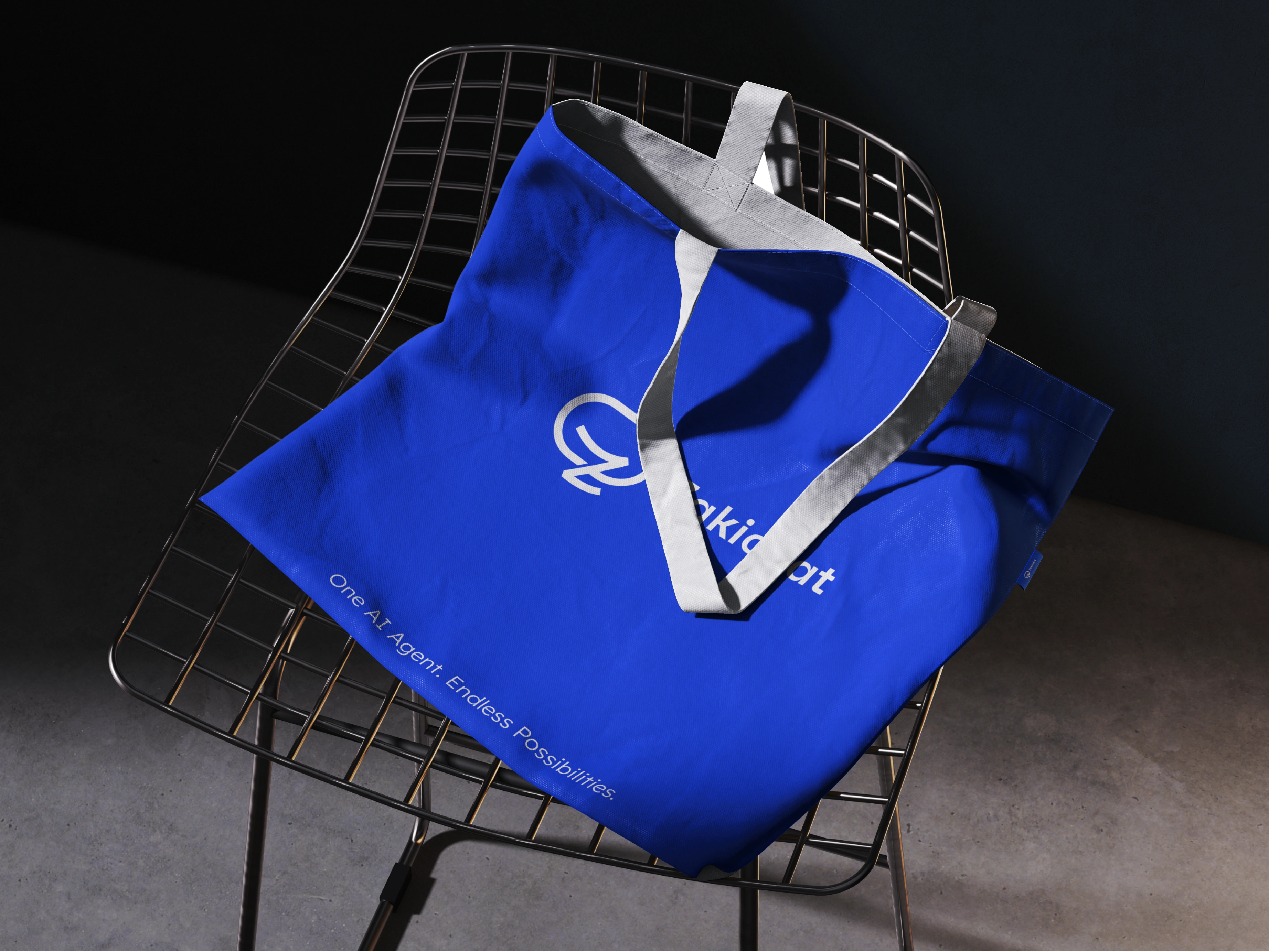 A royal-blue Zakichat tote bag with grey webbing handles draped over a black wire-mesh chair in a dark studio, the handles throwing a hard shadow across the printed mark and wordmark.